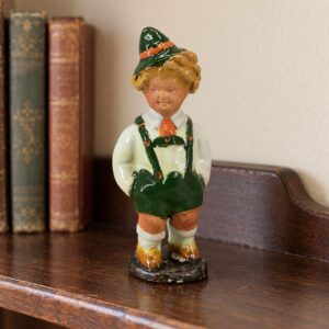 Szécsi Jolán tiroli kisfiút ábrázoló kerámiafigurája könyvek mellett, fa polcon / Szécsi Jolán ceramic Tyrolean boy figurine beside books on a wooden shelf