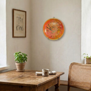 A vibrant orange and yellow glazed studio pottery wall plate with an incised dark abstract sunburst design. Shown hanging on a white wall in a cozy room and in close-up shots revealing the artist's handwritten signature on the back.