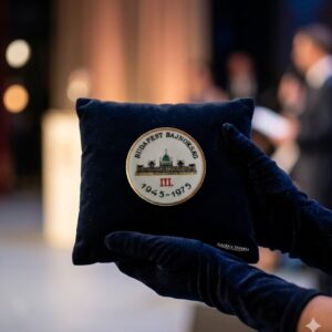 A round white vintage Hollóháza porcelain commemorative plaque with a gold rim, featuring a graphic of the Hungarian Parliament and the text 'BUDAPEST BAJNOKSÁG III. 1945-1975'. Held elegantly on a dark blue velvet pillow in one shot, and showing the green maker's mark on the back in another.