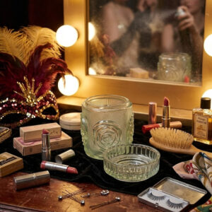 Hollywoodi budoár képe egy Ząbkowice üveg púdertartó tégellyel. A vintage Polish Ząbkowice pressed glass powder jar with a lid, featuring a geometric rose pattern and a slight green tint, displayed beautifully on a wooden vanity table among classic cosmetics, lipsticks, and a mirror.