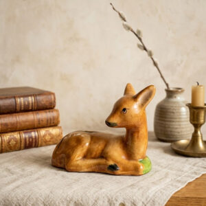 A handcrafted Hungarian ceramic lying fawn figurine from the Bodrogkeresztúr factory, resting on an oatmeal linen surface, next to a small wooden candlestick with a lit beeswax candle, three smaller antique books with aged spines and typography, and a miniature vase with a delicate dried pussy willow and flower. A small metal tag with the number "38/2201" is discreetly integrated into the scene. Soft natural window light.