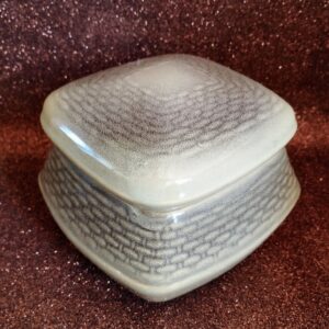 Angled shot of a closed square ceramic bonbonnier, showing the base and lid, on a sparkly brown background.