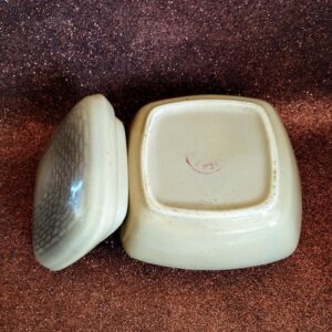 Underside view of the square ceramic bonbonnier base and the separate lid, showing the pink maker's mark, on a sparkly brown background.