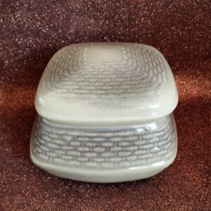 Full shot of a closed square ceramic bonbonnier with a basketweave base and textured glaze lid, on a sparkly brown background.