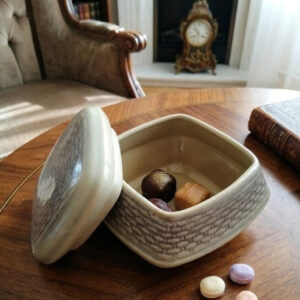 Close-up of a square ceramic bonbonnier, lid open on a wooden table, containing two chocolates and another nearby.